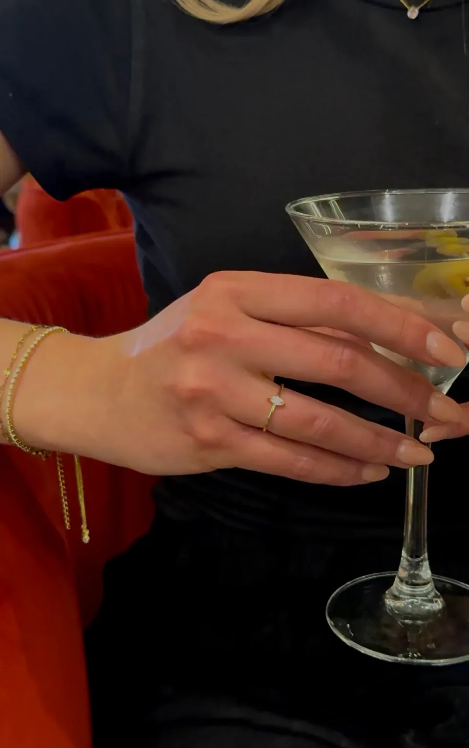 A hand around a martini stem wearing the Astra ring, with gold bracelets stacked at the wrist